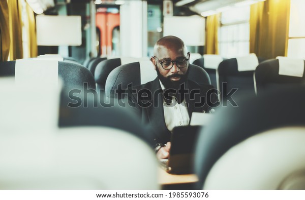 A dapper bearded bald black man entrepreneur in\
eyeglasses is using his laptop while sitting inside of a high-speed\
intercity train surrounded by seats, selective focus, shallow depth\
of field