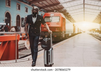 A Dapper Bearded Bald Black Senior Businessman In A Fashionable Suit With A Vest And Bowtie Is Standing On A Platform Of A Railway Station Depot With His Baggage, Next To The Red Railroad Train