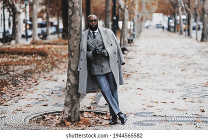 A dapper black senior entrepreneur in eyeglasses and an elegant outfit with a custom-made costume and a coat, is leaning against a tree on an autumn boulevard alleyway; a copy space place on the right