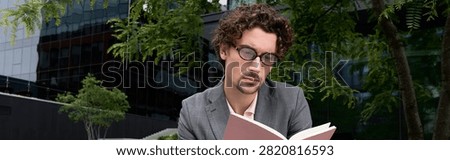 A dapper man with elegant attire sits on the street, engrossed in a book amidst modern buildings.
