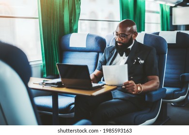 A dapper mature bald bearded black man entrepreneur is using his laptop and holding a document in his hand while sitting on a soft seat of a high-speed intercountry train during his business travel