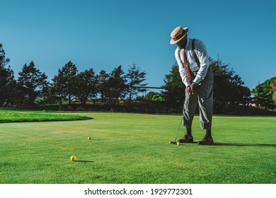 A Dapper Mature Bearded Black Guy In An Elegant Outfit And A Hat, With A Cigar In His Mouth, Is Standing On A Green Lawn Of A Golf Field Is Ready To Hit A Ball Using A Club On A Warm Sunny Evening