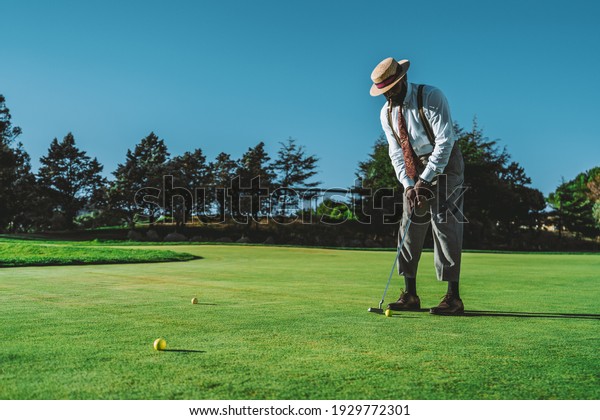 A dapper mature bearded black guy in an elegant\
outfit and a hat, with a cigar in his mouth, is standing on a green\
lawn of a golf field is ready to hit a ball using a club on a warm\
sunny evening