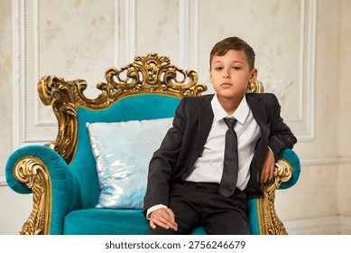 A dapper young boy, dressed in a sharp black suit and tie, sits with a composed demeanor on an elaborate teal velvet armchair.