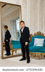 A dapper young boy, wearing a smart navy suit and tie, stands indoors in a grand room during the day.