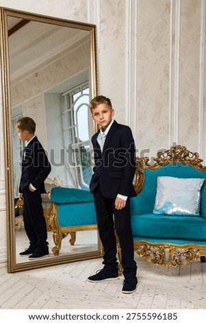 A dapper young boy, wearing a smart navy suit and tie, stands indoors in a grand room during the day.