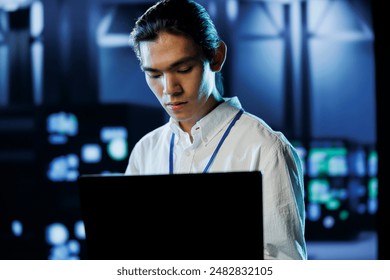 Dara center manager running code and programming on laptop, troubleshooting and upgrading high tech facility designed to accommodate rows of fully operational server supercomputers