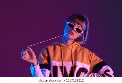 Daring pretty young 20s fashion teen girl model wear glasses chewing bubble gum look at camera stand at purple studio background, igen teenager with bubblegum in trendy night 80s party light portrait - Shutterstock ID 1557182312
