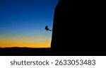 A daring rock climber, silhouetted against a vibrant sunset, defies gravity as they scale a steep cliff. The breathtaking scene showcases the thrill and beauty of extreme sports. Aerial view.