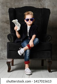 Daring And Sassy Young Rich Boy Kid Millionaire Sits In Big Luxury Armchair And Demonstrates Bundle Of Money Dollar Bills