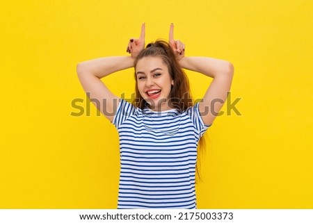 Daring stylish brunette girl in white-blue striped t shirt making confident and amused expression showing horns with index fingers on head. Indoor studio shot on yellow background