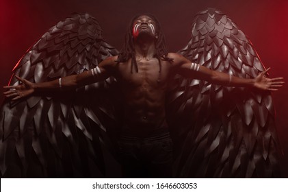 Dark African Angel With Big Black Wings Isolated, Young Serious Muscular Man Wearing Big Wings On The Back. Strong Angel Come Down From Heaven. Fantasy