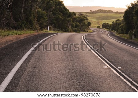 Dark asphalt road with bright white lines curves downhill with trees on sides and green farm fields and horses in far away in bright sunrise in Victoria, Australia
