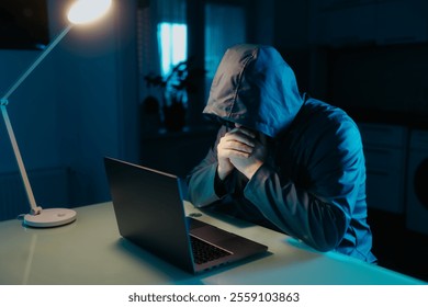Dark atmosphere of a home kitchen, where a man in a hoodie focuses on his laptop, embodying the modern-day challenges of cybersecurity.