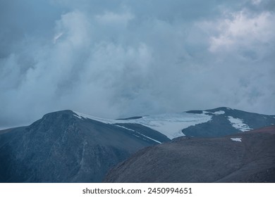 Dark atmospheric alpine landscape with big rocky mountain silhouettes with snow under low clouds in dramatic gray cloudy sky. Large mountain range with glacier on top under low cloudiness at twilight. - Powered by Shutterstock