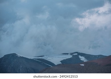 Dark atmospheric alpine landscape with big rocky mountain silhouettes with snow under low clouds in dramatic gray cloudy sky. Large mountain range with glacier on top under low cloudiness at twilight. - Powered by Shutterstock