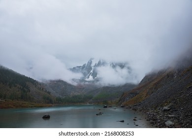 Dark Atmospheric Landscape With Alpine Lake And High Snow Mountains In Dense Fog. Large Snowy Mountain Range In Thick Fog In Dramatic Overcast. Snow-covered Rocky Mountains In Low Clouds During Rain.