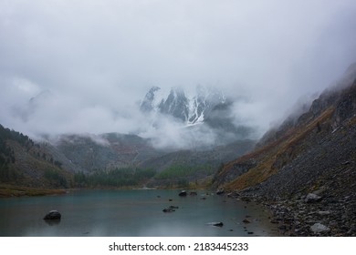 Dark Atmospheric Landscape With Alpine Lake And High Snow Mountains In Dense Fog. Large Snowy Mountain Range In Thick Fog In Dramatic Overcast. Snow-covered Rocky Mountains In Low Clouds During Rain.