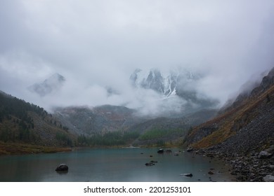 Dark Atmospheric Landscape With Alpine Lake And High Snow Mountains In Dense Fog. Large Snowy Mountain Range In Thick Fog In Dramatic Overcast. Snow-covered Rocky Mountains In Low Clouds During Rain.