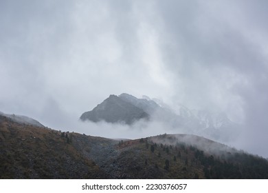 Dark atmospheric landscape with high mountain silhouettes in dense fog in rainy weather. Snowy rocky mountain top above hills in thick fog in dramatic overcast. Black rocks in low clouds during rain. - Powered by Shutterstock