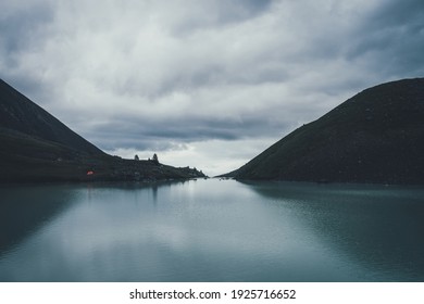 Dark atmospheric landscape with orange tent near mountain lake in highland valley under cloudy sky in dark time. Ripples on mountain lake water in dusk. Dark scenery with water ripple on highland lake - Powered by Shutterstock