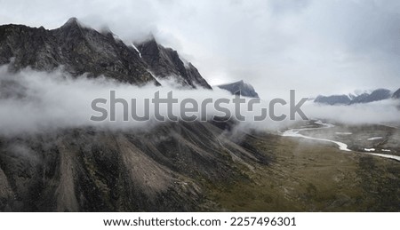 Dark atmospheric surreal landscape with dark rocky mountain top in low clouds in gray cloudy sky. Gray low cloud on high pinnacle. High black rock with snow in low clouds. Surrealist gloomy mountains