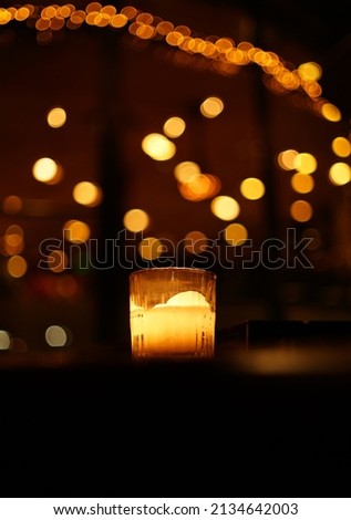 Dark background with burning glass candle and golden bokeh. Candlelight with fairy lights bokeh in front of cocktail bar. One candle flame light at night with bokeh on dark background.
