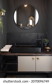 Dark bathroom, black sink, round mirror with black frame, metal wall cladding, gilded square wooden cabinet handles, rolled white towels, brown marble surface, flower pots and accessories