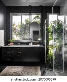 Dark Bathroom, Black Wood Cabinet With Gilt Handles, Oval-shaped Mirror, Hanging From The Ceiling, Black Floor Tiles, Black Walls, Antique Persian Rug, Large Flower Pot, Gilded Faucet And Accessories
