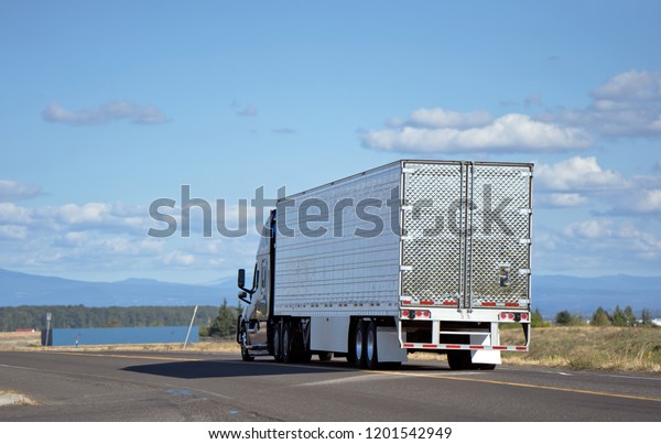Dark big rig modern professional popular semi truck\
transporting perishable food in chilled refrigerated semi trailer\
running on divided local road with river view and clouds for\
delivery goods