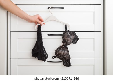 Dark black lace bra and panties hanging on hanger at white drawer. Young adult woman hand taking underwear. Closeup. Front view.