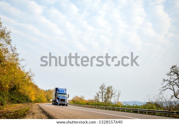 Dark blue bonnet modern American big rig classic\
freight transportation bonnet semi truck with black grille\
transporting commercial cargo in dry van semi trailer on the\
winding autumn local road