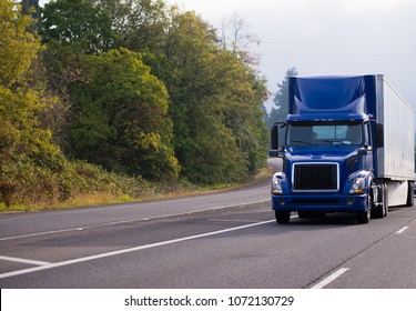 Dark Blue Day Cab Big Rig Semi Truck Fleet For Community Local Deliveries Of Goods To The Stores With Dry Van Semi Trailer Running On Wide Highway With Green And Yellow Trees