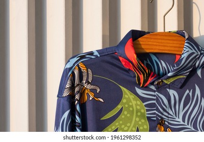 Dark Blue Hawaiian Shirt With Tropical Foliage Pattern On Hanger Hanging On White Wooden Battens Background  