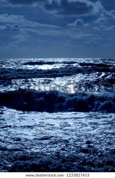 A dark blue sea with waves swelling the sea has\
dark shadows where the waves are swelling and the sunlight is\
glistening white on the sea on the horizon in the foreground is a\
dark pebble beach