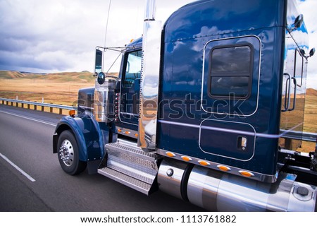 Dark blue shiny classic American big rig semi truck with chrome accessories with reflection on cab surface running on the road with yellow summer field on background in California
