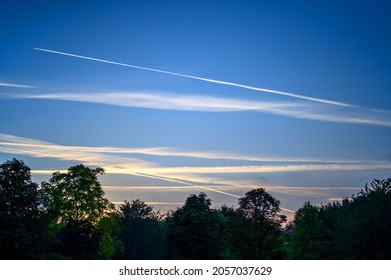 Dark blue sky at dusk with white clouds. An airplane (aeroplane) contrail extends across the sky. Silhouette of trees on the horizon. View of the sky with clouds and contrail just after sunset.