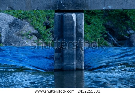 Dark blue water flows fast past a concrete pillar and spills over an edge with green and concrete in the background