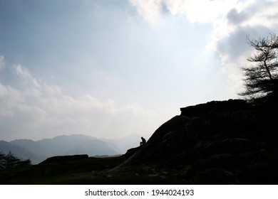 Dark, brooding, dramatic: silhouette of an anonymous figure sat on a mountain top. Sharp against a bright cloudy sky; one tree, and further mountains in the hazy distance.
