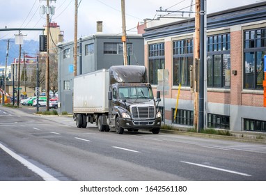 Dark brown Big rig day cab semi truck with roof spoiler transporting goods in short dry van semi trailer for improved maneuverability running uphill on the city street with multilevel buildings