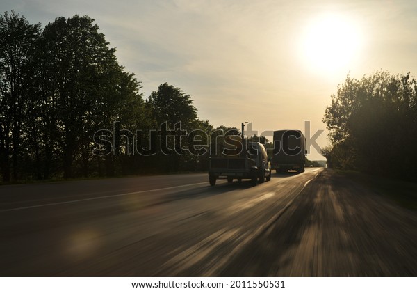Dark car with the\
trailer and a truck on the countryside road in motion with trees\
against sky with bright\
sun
