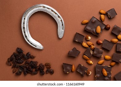 Dark Chocolate Pieces With Roasted Almonds And Raisins Scattered Around Horse Shoe On Brown Background