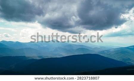 Dark cloud bases hover above a sea of blue mountains where narrow valleys gleam with light. The fore ridge lies in shadow giving depth and mood to this airy and remote terrain