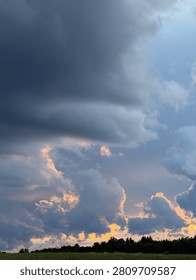 Dark clouds fill the sky over a green field. The setting sun shines through the clouds, creating shades of orange and blue in the sky.