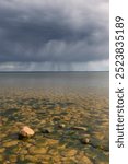 Dark clouds loom over Lake Vanern, casting shadows on the tranquil waters and rocky shoreline. The atmosphere suggests an approaching storm, enhancing the serene beauty.