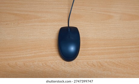 A dark computer mouse with a wire rests on a wooden surface. Technology and office equipment.