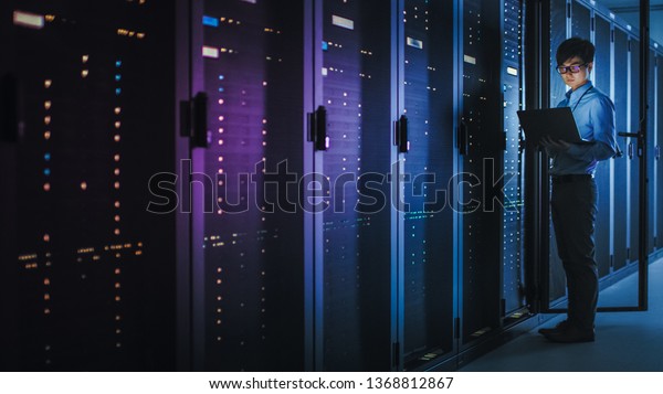 In Dark Data Center: Male IT Specialist Stands\
Beside the Row of Operational Server Racks, Uses Laptop for\
Maintenance. Concept for Cloud Computing, Artificial Intelligence,\
Supercomputer. Neon Lights
