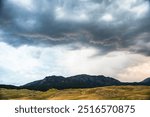 Dark, dramatic clouds gather over rolling hills and distant mountains.