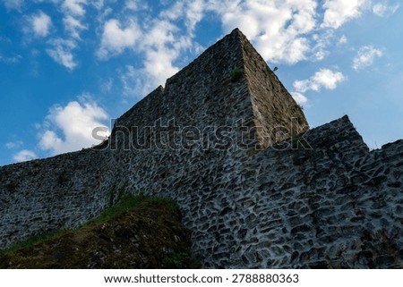 Dark dramatic silhouette of towering medieval stone castle walls reaching up towards a bright cloudy sky.