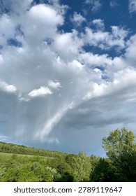 Dark dramatic storm clouds hanging low over a green forest landscape before rainfall. Powerful sky with heavy rain shaft, contrasting with a strip of blue sky near the horizon. Moody weather scene in nature, summer thunderstorm approaching.